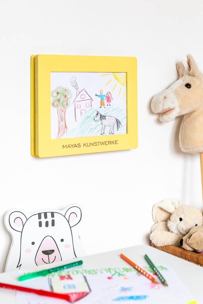 May include: A yellow framed drawing of a house, tree, people, and a horse, with the text "MAYAS KUNSTWERKE". A stuffed horse head and a bear-shaped decoration are also visible.