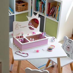 Könnte beinhalten: Ein rosa-weißes Holz-Schminktisch-Set mit einem Spiegel, Schubladen und Fächern zur Aufbewahrung von Make-up und Accessoires. Der Schminktisch hat ein Kronendesign und der Text "Lea's Beautification" ist auf der Vorderseite eingraviert. Der Schminktisch steht auf einem weißen Tisch mit zwei weißen Stühlen mit Bärenohren.