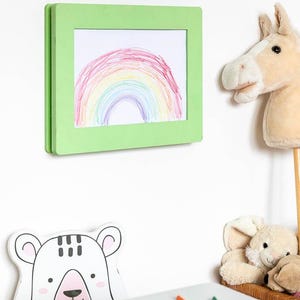 May include: A green framed drawing of a rainbow hangs on a white wall. A plush horse head on a stick and a stuffed bunny are nearby. A white bear-shaped cutout and colored pencils are on a table.