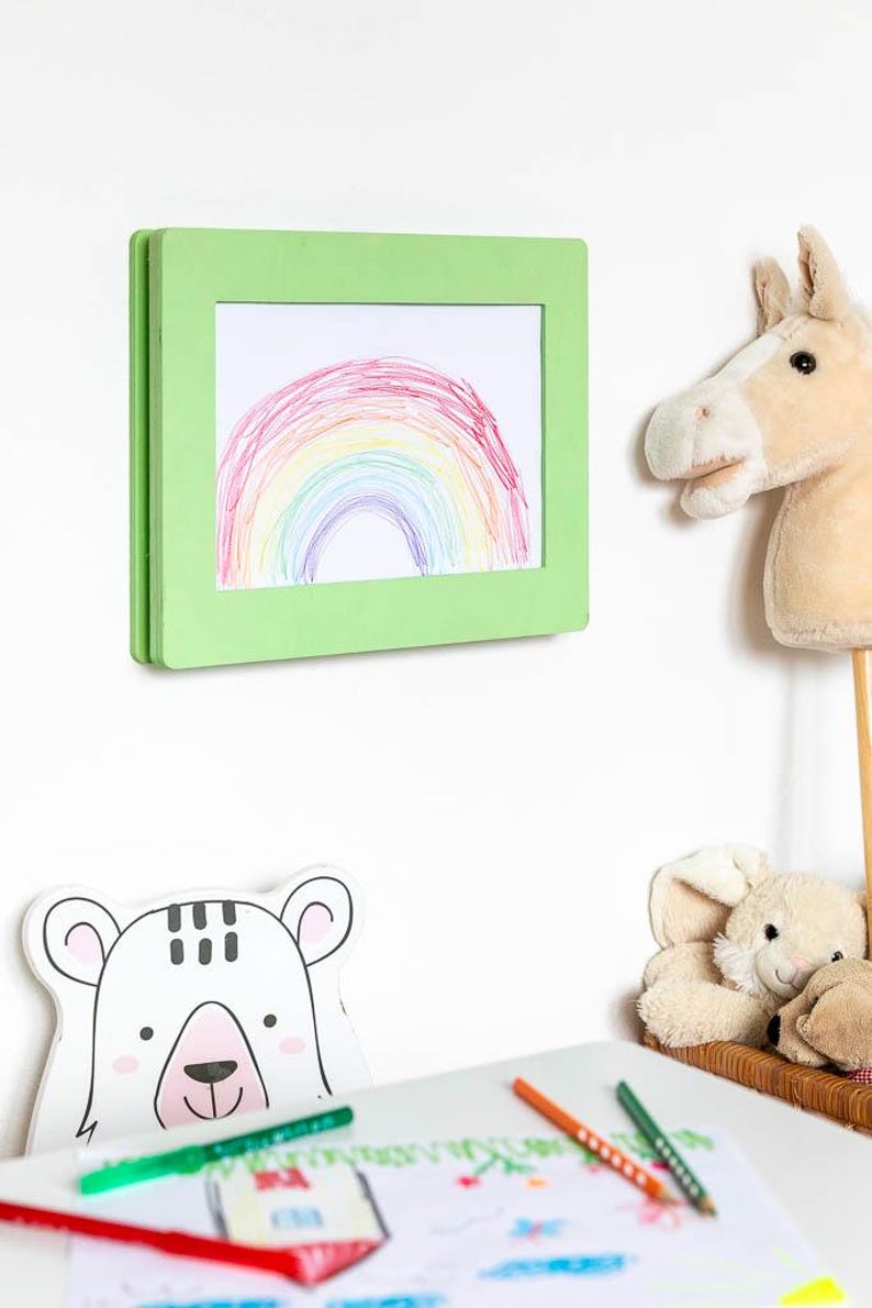 May include: A green framed drawing of a rainbow hangs on a white wall. A plush horse head on a stick and a stuffed bunny are nearby. A white bear-shaped cutout and colored pencils are on a table.
