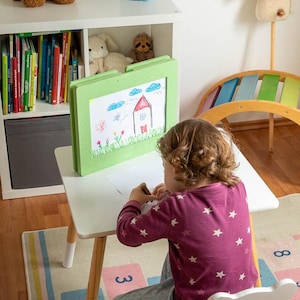 Könnte beinhalten: Eine Kinderzeichnung auf einer grün gerahmten Staffelei auf einem weißen Tisch. Die Zeichnung zeigt ein Haus, Blumen und Schmetterlinge. Ein brauner Teddybär, ein blauer Lautsprecher und Stofftiere befinden sich auf einem weißen Regal.