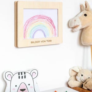 May include: A wooden frame displaying a child's drawing of a rainbow in red, orange, yellow, green, blue, and purple. The text "BILDER VON TOBI" is printed below the drawing. A stuffed horse head and a bear-shaped drawing are also visible.