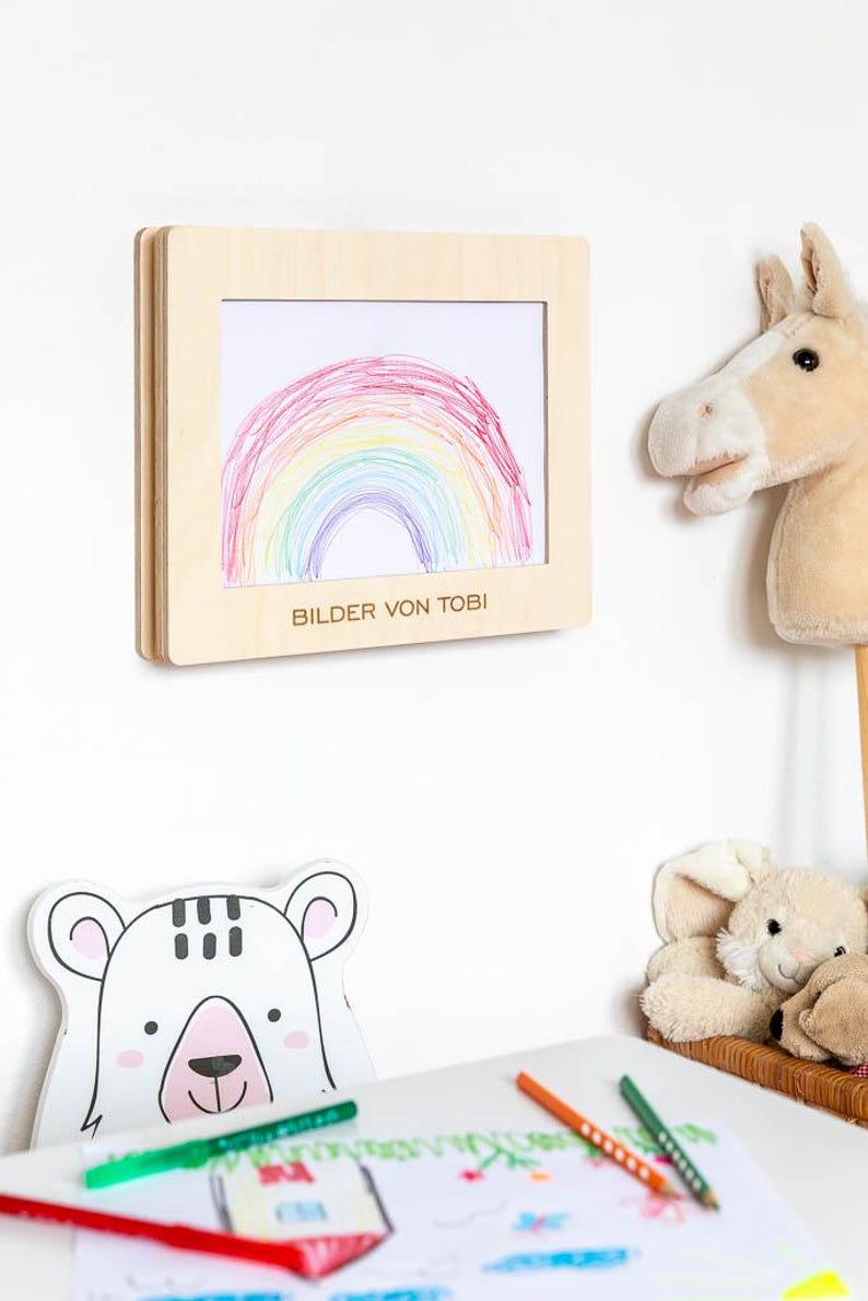 May include: A wooden frame displaying a child's drawing of a rainbow in red, orange, yellow, green, blue, and purple. The text "BILDER VON TOBI" is printed below the drawing. A stuffed horse head and a bear-shaped drawing are also visible.