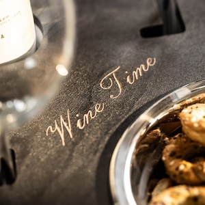 May include: A black slate serving tray with the words "Wine Time" engraved in gold lettering. The tray has a built-in glass holder and a small bowl filled with pretzels.
