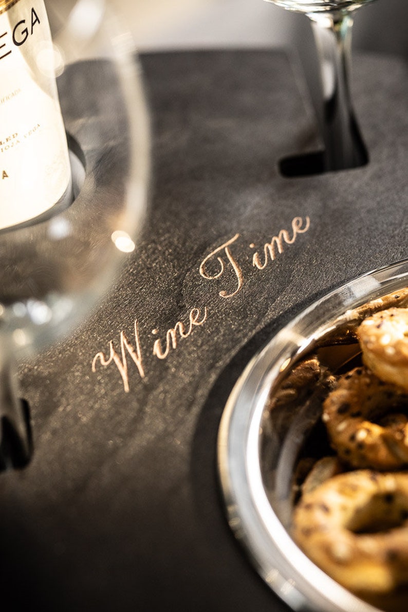 May include: A black slate serving tray with the words "Wine Time" engraved in gold lettering. The tray has a built-in glass holder and a small bowl filled with pretzels.