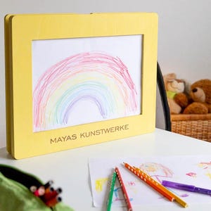 May include: A yellow framed artwork featuring a colorful rainbow drawing. The frame has the text "MAYAS KUNSTWERKE" on the bottom. Colored pencils and other drawings are on the table. A green pencil case is in the foreground.