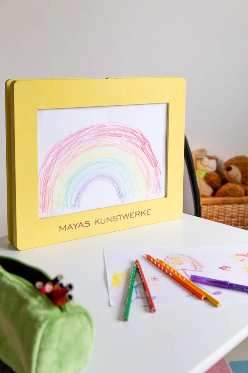 May include: A yellow framed artwork featuring a colorful rainbow drawing. The frame has the text "MAYAS KUNSTWERKE" on the bottom. Colored pencils and other drawings are on the table. A green pencil case is in the foreground.