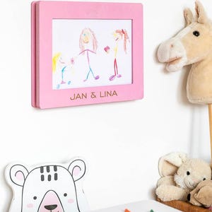 May include: A pink wooden frame displaying a child's drawing of three figures, with the text "JAN & LINA" at the bottom. A plush horse head and a bear-shaped decoration are also visible. The drawing is in crayon.