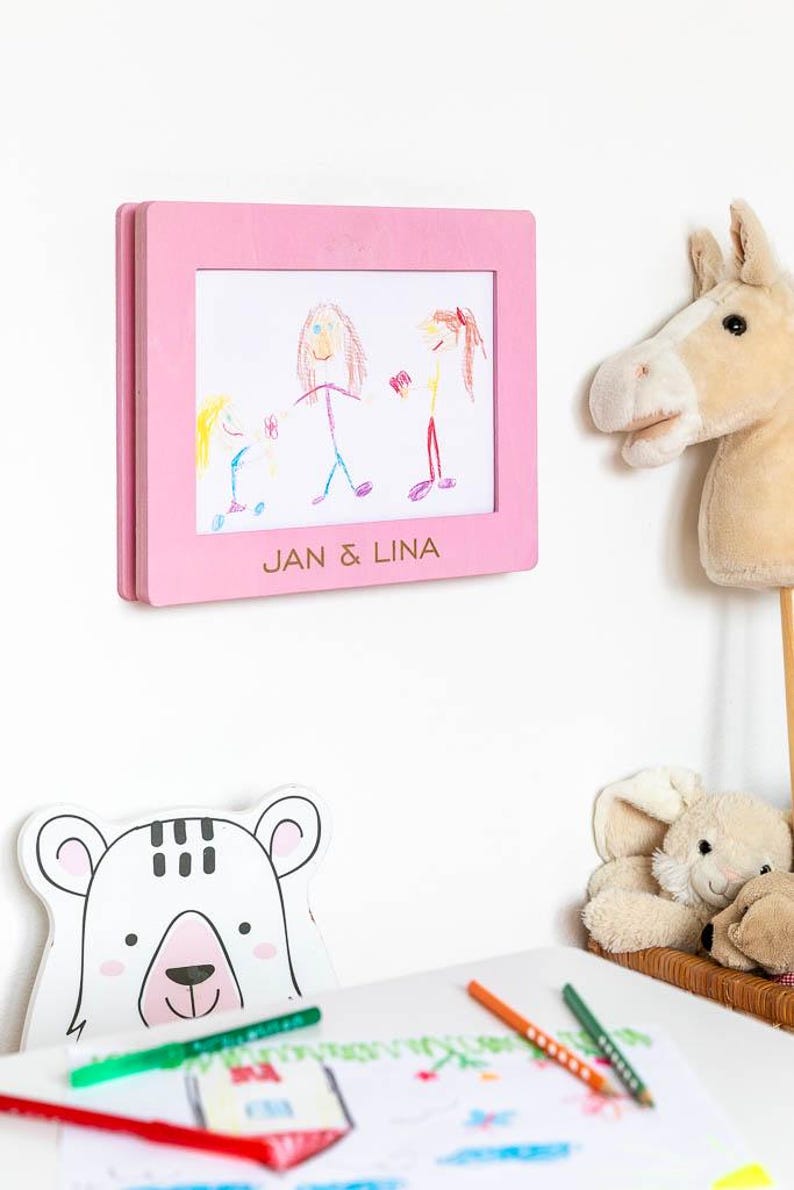 May include: A pink wooden frame displaying a child's drawing of three figures, with the text "JAN & LINA" at the bottom. A plush horse head and a bear-shaped decoration are also visible. The drawing is in crayon.