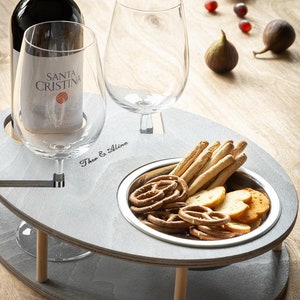 May include: A gray wooden serving tray with two wine glass holders, a bottle holder, and a small bowl for snacks. The tray is engraved with the words "Theo & Alina".