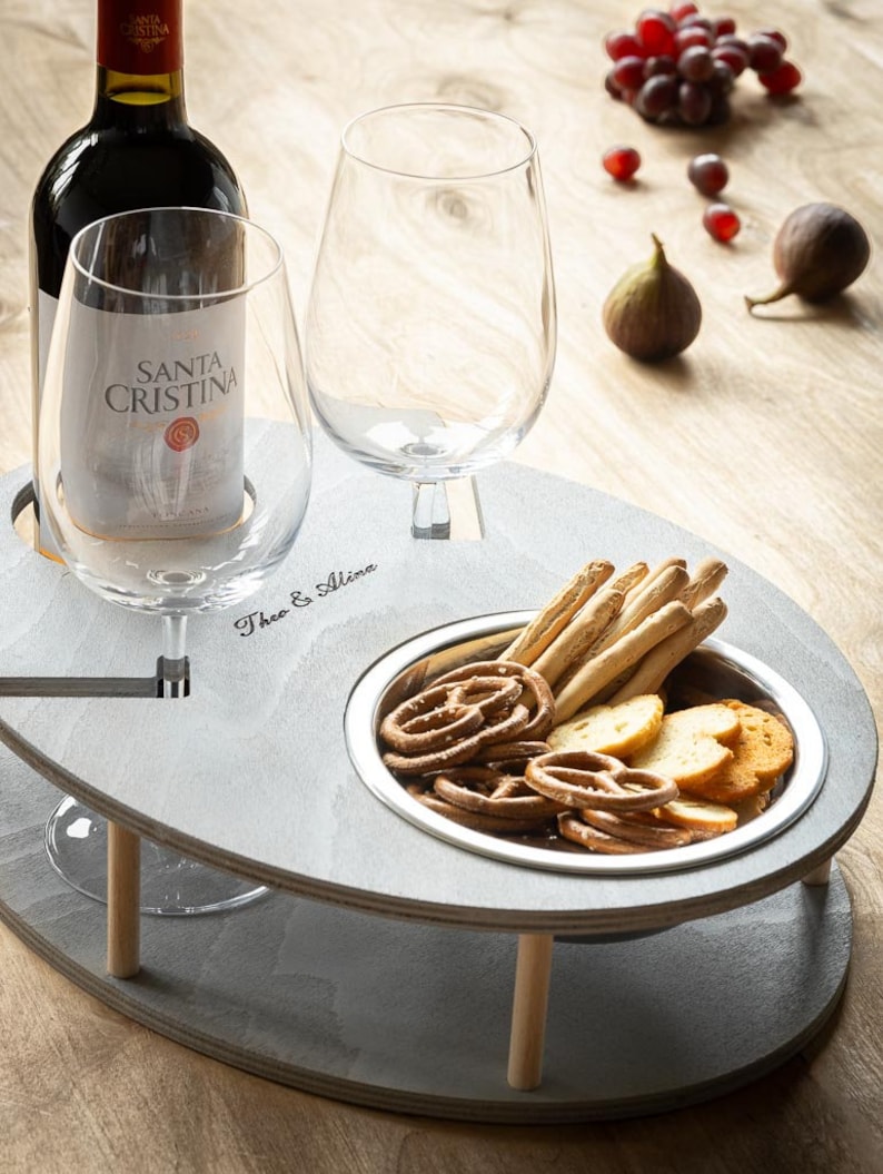 May include: A gray wooden serving tray with two wine glass holders, a bottle holder, and a small bowl for snacks. The tray is engraved with the words "Theo & Alina".