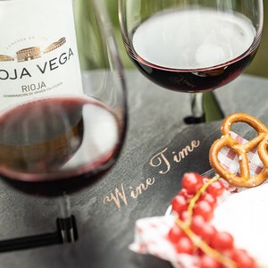 May include: A black slate serving tray with the words "Wine Time" inscribed in gold lettering. The tray has two glasses of red wine, a pretzel, and red currants on a white napkin.