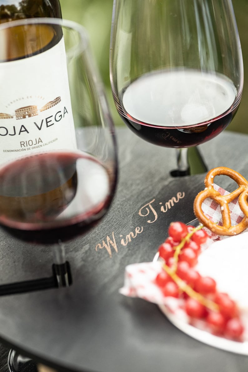 May include: A black slate serving tray with the words "Wine Time" inscribed in gold lettering. The tray has two glasses of red wine, a pretzel, and red currants on a white napkin.