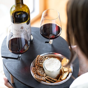May include: A black wooden serving tray with two wine glasses, a bottle of wine, and a small bowl of crackers and dip. The tray has two wooden legs and is designed to hold wine glasses securely.