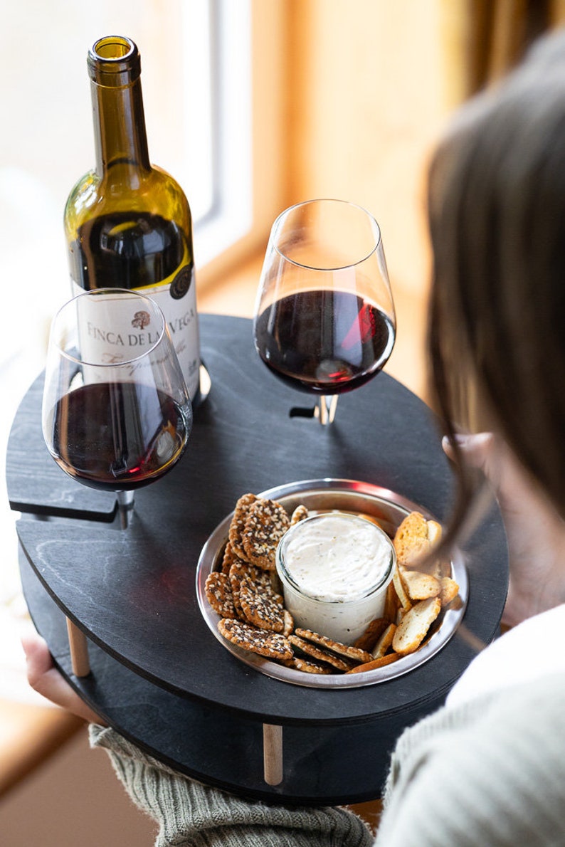 May include: A black wooden serving tray with two wine glasses, a bottle of wine, and a small bowl of crackers and dip. The tray has two wooden legs and is designed to hold wine glasses securely.