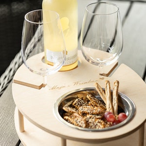 May include: A wooden serving tray with two wine glass holders, a bowl for snacks, and the text "Maja's Weinbar" engraved on the surface. The tray is holding a bottle of white wine, a bowl of crackers, and grapes.