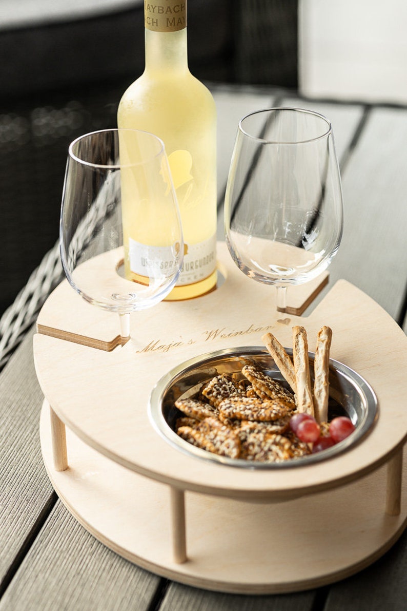 May include: A wooden serving tray with two wine glass holders, a bowl for snacks, and the text "Maja's Weinbar" engraved on the surface. The tray is holding a bottle of white wine, a bowl of crackers, and grapes.