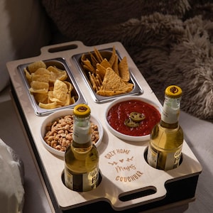 Könnte beinhalten: Ein Holztablett mit vier Fächern für Snacks und zwei Schlitzen für Getränke. Das Tablett hat eine schwarze Basis und ist mit dem Text "Cozy Home Maya's Couchbar" graviert. Das Tablett ist mit Snacks gefüllt, darunter Chips, Erdnüsse und Salsa.