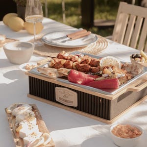 Könnte beinhalten: Ein Serviertablett aus Holz mit einer Metalloberfläche enthält gegrillte Hähnchenspieße, rote Paprika und Fladenbrot. Das Tablett steht auf einer weißen Tischdecke, mit Zitronen, einem Weinglas und einer Schüssel mit Dip in der Nähe. Das Tablett trägt den Text "HOTTY JAN".