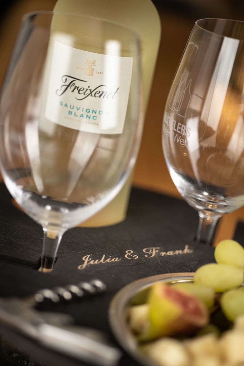 May include: A black slate serving tray with the words "Julia & Frank" engraved on it. A bottle of Freixenet Sauvignon Blanc wine is on the tray, along with two wine glasses, a corkscrew, and a bowl of fruit.