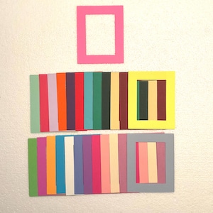 May include: A collection of colorful paper frames and strips arranged on a white surface. The frames are in various colors, including pink, yellow, and gray. The strips are arranged in rows, showcasing a spectrum of hues.