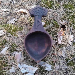 Op de afbeelding: Een donkerbruine houten lepel met een gegraveerd handvat met een knoopwerkontwerp. De lepel ligt op een bed van mos en dennennaalden.