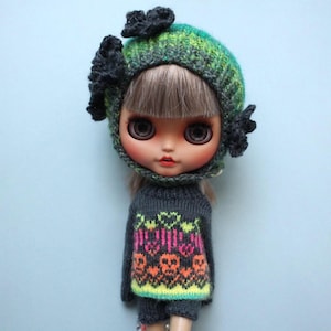 May include: A doll wearing a knitted outfit, including a green and black hat with floral accents and a gray sweater with a colorful skull and heart design. The doll's face has large eyes and a neutral expression.