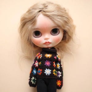 May include: A doll with blonde hair and large eyes. The doll is dressed in a black turtleneck sweater adorned with colourful floral crochet patches. The doll's face has a soft, blushed appearance, with freckles and a gentle expression.
