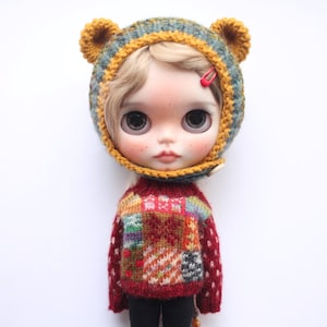 May include: A doll wearing a handmade knitted bear hat with yellow ears and a red hair clip. The doll is also wearing a red sweater with a patchwork design and black trousers. The doll has large eyes and rosy cheeks.