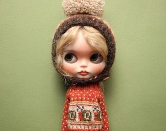 Puede incluir: Una muñeca de cabello rubio que lleva un gorro de punto marrón oscuro rematado con un gran pompón beige. La muñeca viste un suéter naranja de manga larga con un patrón repetitivo de pequeños caracoles en el pecho y pantalones de color verde oscuro.