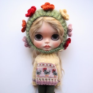 May include: A doll wearing a light green knitted hat decorated with colourful flower accents. The doll is also wearing a yellow sweater with a pink and white pattern featuring three chickens. The doll has blonde hair and large eyes.