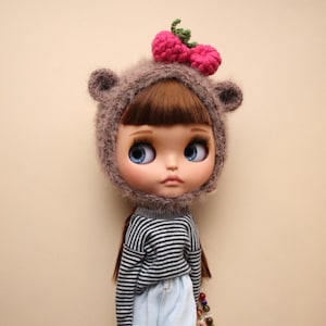 May include: A doll wearing a brown bear hat with pink crochet cherries and a gray and white striped sweater. The doll has large blue eyes and is wearing light blue jeans. The hat has small bear ears.
