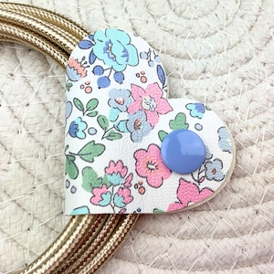 Peut inclure: Un organisateur de cordon en forme de cœur avec une base blanche ornée d'un motif floral dans des tons rose, bleu clair et vert. Un bouton-pression bleu pervenche uni sécurise le pli, tandis qu'un cordon doré métallique est enroulé dessous.