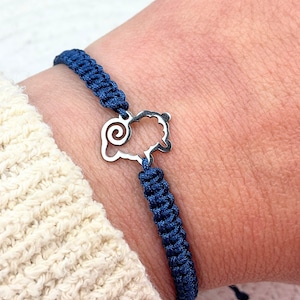 Könnte beinhalten: Ein Armband aus dunkelblauem geflochtenem Kordelmaterial mit einem silberfarbenen Metallanhänger in Form eines stilisierten Schafs mit einem markanten Spiralhorn. Die Kordel ist in einem engen, sich wiederholenden Knotenmuster gewebt.