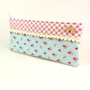 Könnte beinhalten: Rechteckiges Federmäppchen mit hellblauem Blumenmuster auf der unteren Hälfte und einem rosa-weiß karierten Muster auf der oberen Hälfte. Eine cremefarbene Pomponborte trennt die beiden Muster. Oben befindet sich ein kleiner Holzknopf in Form einer Schleife.