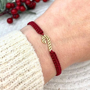 Könnte beinhalten: Ein rotes geflochtenes Armband mit einem goldfarbenen Zuckerstangen-Anhänger. Das Armband wird am Handgelenk getragen, wobei der Zuckerstangen-Anhänger im Mittelpunkt steht. Ein festliches Accessoire.