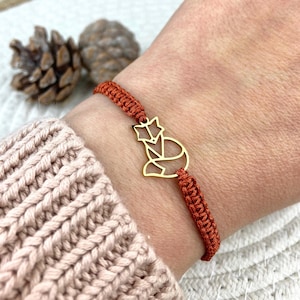 Könnte beinhalten: Armband mit einem goldfarbenen Fuchs-Anhänger und einem gewebten, rostfarbenen Band. Das Armband wird am Handgelenk getragen, im Hintergrund ein hellrosa Pullover. Im Hintergrund sind zwei Tannenzapfen zu sehen.