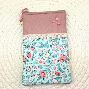 Könnte beinhalten: Ein rechteckiger Beutel mit rosa Oberteil und floral gemustertem Boden. Der Boden hat einen hellblauen Hintergrund mit rosa Blumen, Erdbeeren und Vögeln. Die Oberseite hat drei rosa herzförmige Knöpfe und eine Spitzenborte.