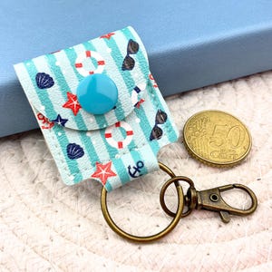 May include: A small, rectangular, light blue and white striped pouch keychain with a snap closure. The fabric features a nautical print of red starfish, white life preservers, dark blue seashells, anchors, and sunglasses. A 50 euro cent coin is shown.