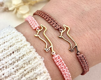 May include: Two braided bracelets with gold-colored dachshund dog charms. One bracelet is pink, the other is brown. The bracelets are worn on a wrist, with a white sweater in the background.