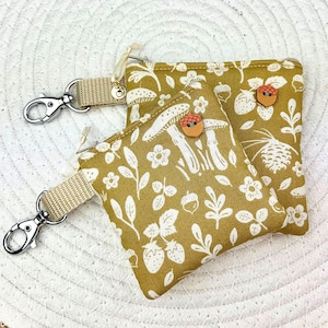 Op de afbeelding: Twee kleine, mosterdkleurige zakjes met een wit bloemen- en paddenstoelenpatroon. Elk zakje heeft een beige bandje met een zilveren sluiting en een houten eikelknop. De zakjes zijn ontworpen voor sleutels of kleine items.