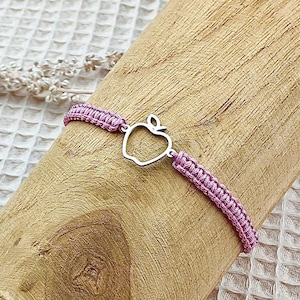 Op de afbeelding: Een roze gevlochten armband met een zilveren appelbedel. De armband ligt op een houten oppervlak.