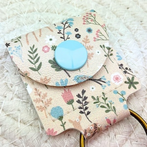 Puede incluir: Un monedero con llavero con estampado floral beige con cierre de botón azul claro. La cartera presenta un llavero dorado y está decorada con patrones florales y de hojas rosas, azules y verdes. La cartera está hecha de un material suave y texturizado.