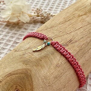 May include: A pink macrame bracelet with a gold feather charm and a small turquoise bead. The bracelet is displayed on a piece of wood, showcasing its woven texture and bohemian style.
