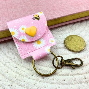 May include: A pink faux leather keychain pouch featuring a white daisy and yellow bee pattern, secured with a yellow heart-shaped snap button. The pouch includes a bronze-toned metal split ring and a lobster clasp, positioned next to a 50 cent coin.
