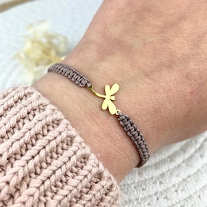 Könnte beinhalten: Taupefarbenes Makramee-Armband mit einem goldfarbenen Libellen-Anhänger. Das Armband besteht aus gewebtem Material. Der Libellen-Anhänger hat ein schlichtes, elegantes Design. Das Armband wird am Handgelenk getragen.