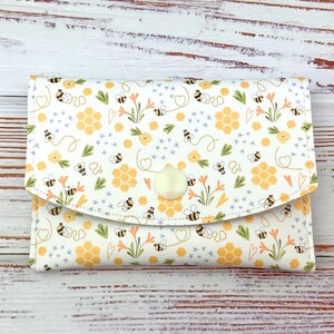 Op de afbeelding: Een witte portemonnee met een klepsluiting, versierd met een patroon van bijen, honingraten, bloemen en harten. Het ontwerp heeft gele, oranje en groene accenten. De portemonnee is vastgezet met een ronde, crèmekleurige knoop.