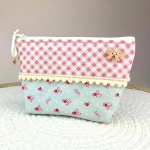 Op de afbeelding: Kleine, rechthoekige stoffen etui met ritssluiting. De bovenkant heeft een roze en wit geruit patroon, de onderkant een bloemenprint op een lichtblauwe achtergrond. Een houten strikvormige knoop en crèmekleurige pompom-bies geven details. Afmetingen: 18cm x 12cm.