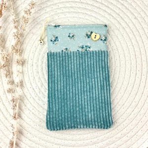 Op de afbeelding: Een teal corduroy telefoonhoesje met een bloemenpatroon. Het bovenste gedeelte heeft een lichtblauwe achtergrond met kleine witte en blauwe bloemen. Een houten hartvormige knoop is aan de bovenkant genaaid.