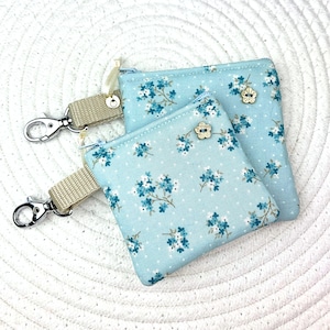 May include: Two small, light blue floral print coin purses with matching keychains. The purses feature a delicate pattern of blue flowers and white polka dots. Each purse has a zipper closure and a decorative flower button.